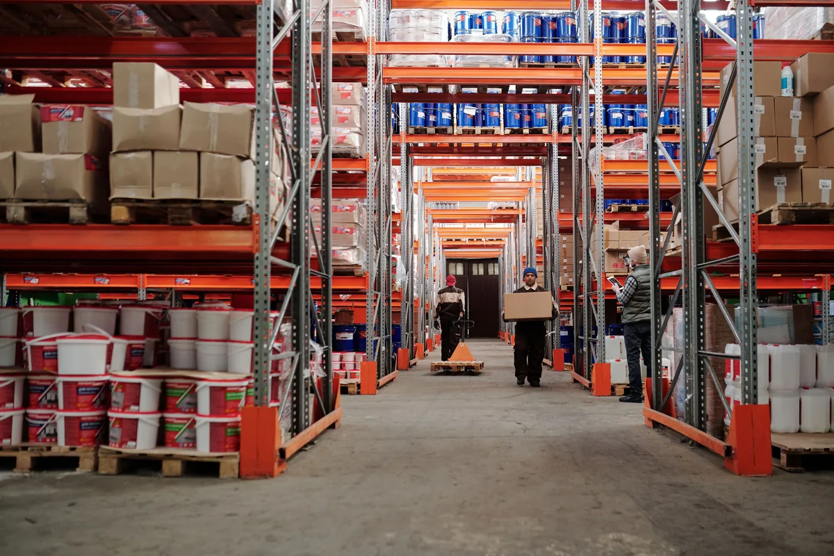 Worker carrying a cardboard box through a warehouse storage aisle.